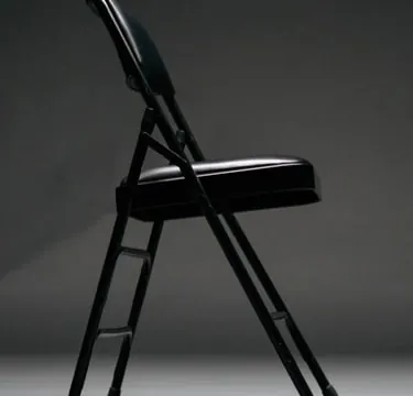 A black folding chair picked out by a single light against a dark studio backdrop.